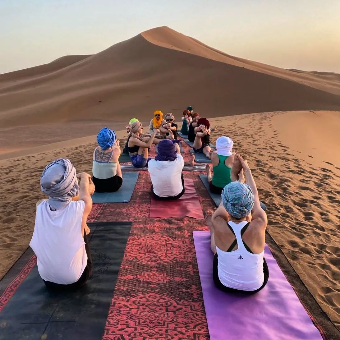 Séance de yoga au lever du soleil face à une grande dune, participants en turbans colorés sur des tapis