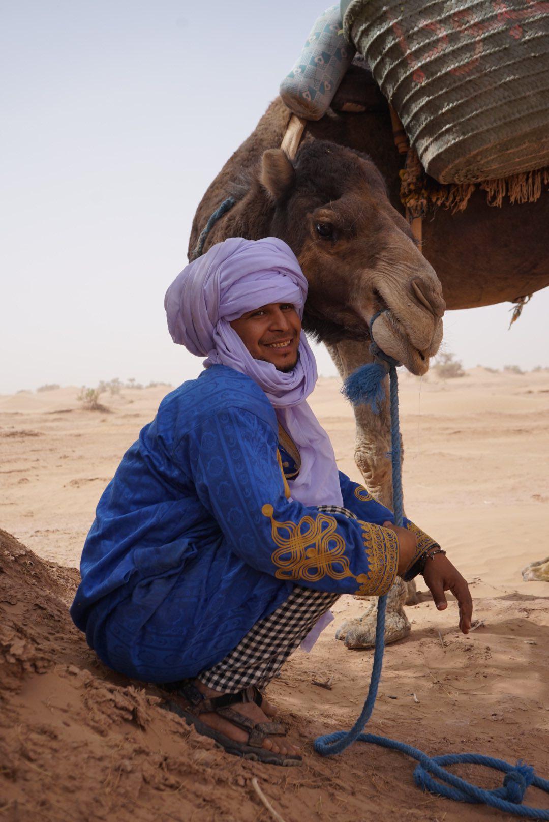 Portrait d'Hassan, guide nomade, accroupi près de son dromadaire, turban lilas